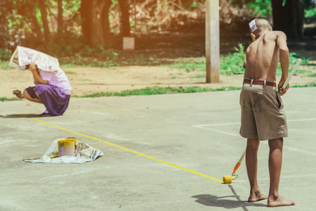 Male students use small paint rollers to paint yellow lines on the cement floor for Sepak Takraw training.の写真素材