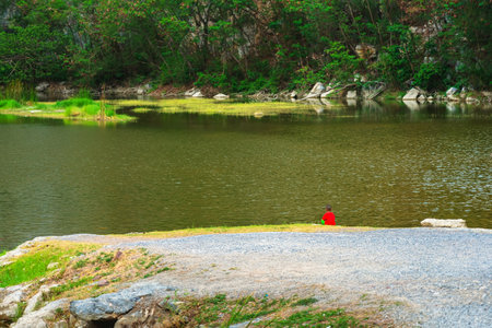 Back view of little boy relaxing alone by the lake with beautiful canyon in background. Boy sits on stone alone at riverside with cliffs and beautiful scenery. Relax with beautiful outdoor natural.の写真素材