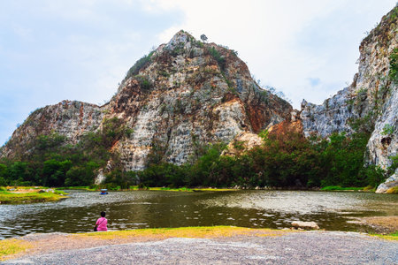 Back view of Asian woman relaxing alone by the lake with beautiful canyon in background. Boy sits on stone alone at riverside with cliffs and beautiful scenery. Relax with beautiful outdoor natural.の写真素材