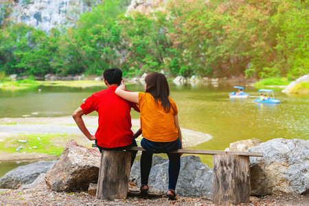Back view of couple relaxing together by the lake with blurred image of happy people enjoying pedal boats or water bikes in lagoon with beautiful canyon in background. Happy family holidays outdoors.の写真素材