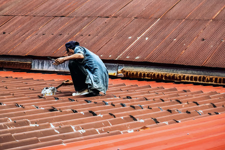 Technician man using polyurethane to seal leak on tiles roof. Man repaired roof by applying sealant to cover rain leaks. Worker man using waterproof roof coating repair to fix crack of old tile roof.の写真素材