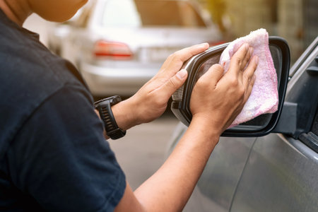 Hands of guy wipes car wing mirror with dry cloth. Pink rag clean car side mirror.  Hand wipes car side mirror. Cleaning wing mirror of auto outdoors. Cleaning retractable rear view lateral car mirrorの写真素材