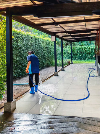 Female janitor cleaning by spraying water and scrubbing walkway. Woman in uniform using water hose flush water to wash floor. Cleaning dirty paving tiles with water hose.の写真素材