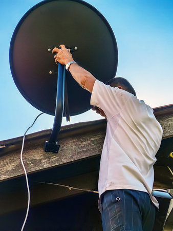 Asian technician installing satellite dish on roof house outdoors. Skillful worker installing satellite dish and television antenna on roof top. Elderly man fitting TV satellite dish to wooden house.の写真素材