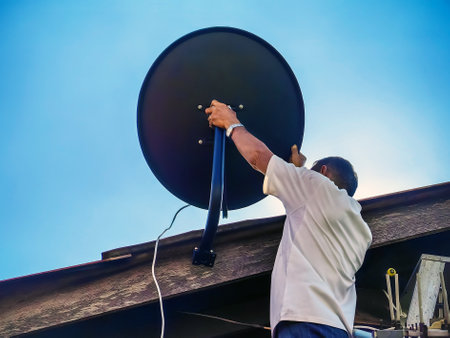Asian technician installing satellite dish on roof house outdoors. Skillful worker installing satellite dish and television antenna on roof top. Elderly man fitting TV satellite dish to wooden house.の写真素材