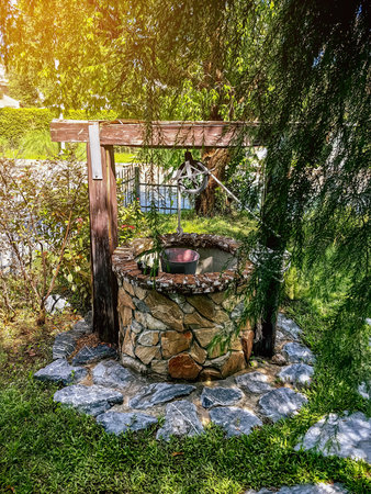 Beautiful artesian well made by stones and wheel pulley with metal bucket and rope in peaceful garden atmosphere. Retro stone water well in rural area. Garden decoration with antique items.の写真素材