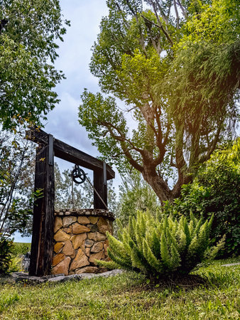 Beautiful artesian well made by stones and wheel pulley with metal bucket and rope in peaceful garden atmosphere. Retro stone water well in rural area. Garden decoration with antique items.の写真素材
