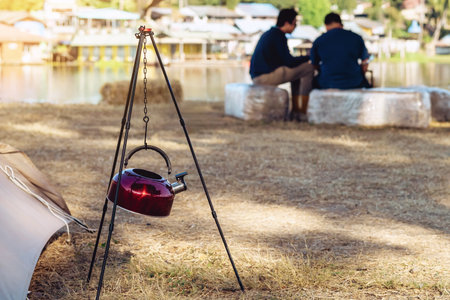 Red stainless steel kettle hang with chain on tripod in camping area. Coffee maker and tent. Insulated hot drink container. Equipment for making portable coffee. Outdoor coffee camping travel vacationの写真素材