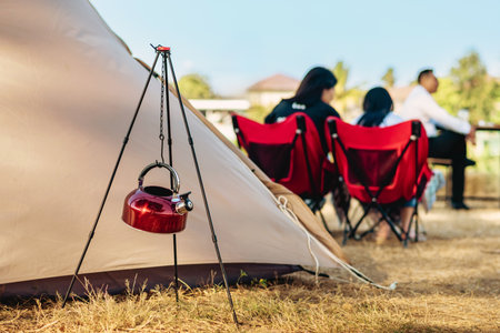 Red stainless steel kettle hang with chain on tripod in camping area. Coffee maker and tent. Insulated hot drink container. Equipment for making portable coffee. Outdoor coffee camping travel vacationの写真素材