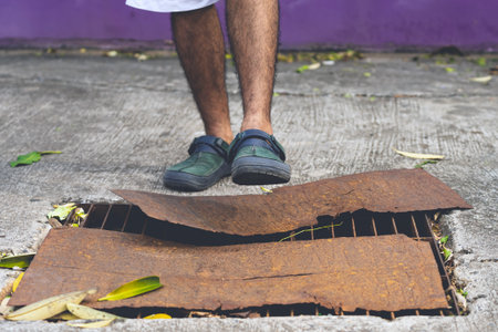 Pedestrian feet wear sandals walk over damaged drain pipe and covered with rusty steel plates on street. Broken manhole. Pothole on road.  Rusty manhole frame. Accident with sewer hatch in city.の写真素材