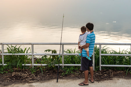 Father with son stand on pier and catching fish on lake background. Family fishing together on pond....