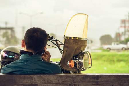 Back view of motorcyclist rests at roadside pavilion and uses smartphone while waiting for rain to stop from heavy rain on side of road. Using communication technology while experiencing travel issuesの写真素材