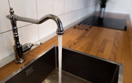 Wasting water by leaving a chrome faucet tap running. Water flowing out of a kitchen stainless steel tap into the sink. Water misuse in domestic duties and activities. Overusing household water.の写真素材
