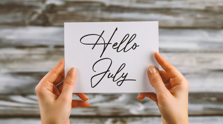 Hands holding a card with the word Hello July on wooden backgroundの素材