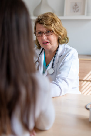 A senior female doctor in a white coat and stethoscope providing a home care consultation to a...