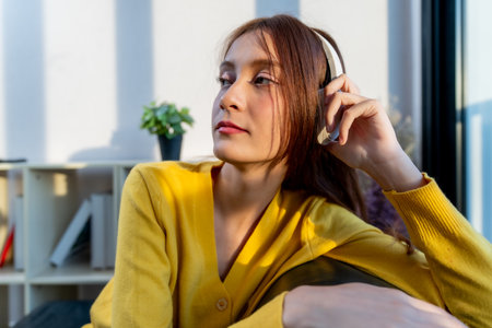 A young woman wearing headphones relaxes in cozy setting, enjoying music. warm sunlight highlights her thoughtful expression, creating serene atmosphereの写真素材