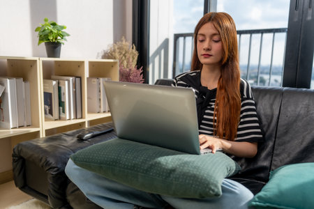 A woman works on laptop while sitting on couch in modern living room. space features natural light, plants, and stylish decor, creating cozy atmosphereの写真素材