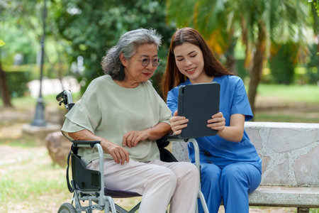 Caring caregiver provides assistance and support to senior in wheelchair while using tablet outdoors, showcasing importance of companionship in careの写真素材
