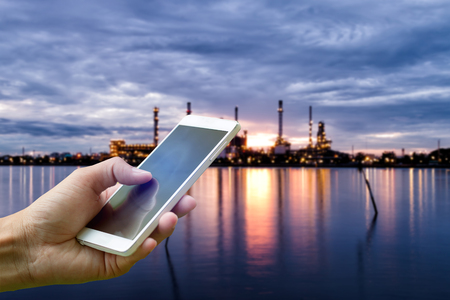 Hand holding smartphone on blurred Oil refinery industry plant along twilight morningの写真素材