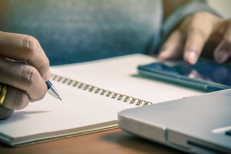 Woman hand writing in notebook with using mobile phone on a desk at home or office.の写真素材