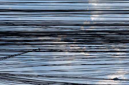 Chaotic mess of electric wires hanged on a pole toward blue sky and cloudsの写真素材