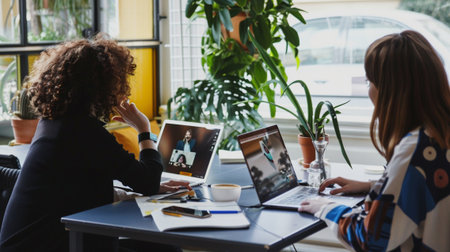 Two women engage in a video call while working on laptops in a vibrant office environment. Green plants add a refreshing touch, emphasizing collaboration and innovation.の素材