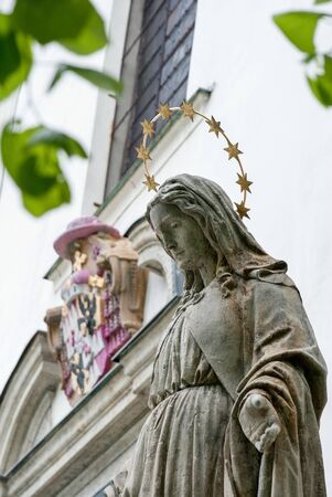 Stone statue of Mary Virgin infront of the white church with green leafs in foregroundの写真素材