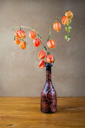 Autumn awesome still life with Physalis alkekengi in decorated bottle placed on wooden board in front of brown wall with clay plasterの写真素材