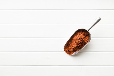 Dark cocoa powder in metal scoop on white table. Top view.