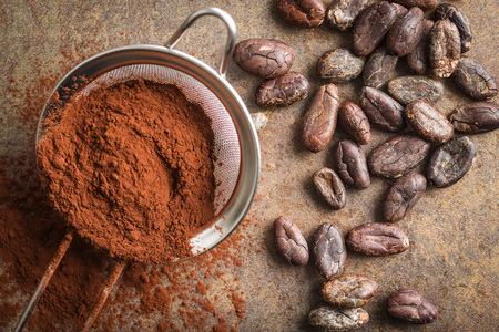 Dark cocoa powder in a sieve and cocoa beans on old kitchen table. Top view.の写真素材