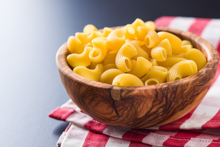 Uncooked elbow macaroni in bowl on checkered napkin.