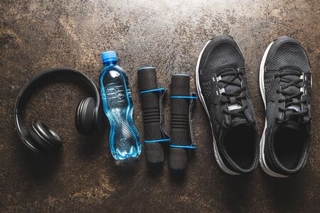 Fitness concept. Black sports shoe, headphsones, dumbbell and bottle of water on old table. Top...