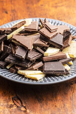 Broken white, milk and dark chocolate bars on plate on wooden table.