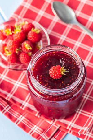 Red rasberries jam in jar and ripe raspberries on checkered napkin.