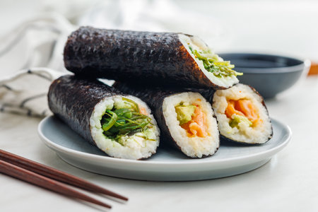 A plate filled with a variety of sushi rolls with a set of wooden chopsticks and a bowl of soy sauce, placed on a simple kitchen table.の写真素材