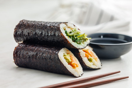 A plate filled with a variety of sushi rolls with a set of wooden chopsticks and a bowl of soy sauce, placed on a simple kitchen table.の写真素材