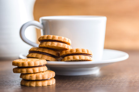 A close-up image of a stack of chocolate-filled cookies resting on a wooden table. A white cup and pitcher are visible in the background, out of focus. The cookies are golden brown in color, with a visible chocolate filling between each layer.の写真素材