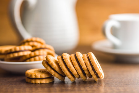A close-up image of a stack of chocolate-filled cookies resting on a wooden table. A white cup and pitcher are visible in the background, out of focus. The cookies are golden brown in color, with a visible chocolate filling between each layer.の写真素材