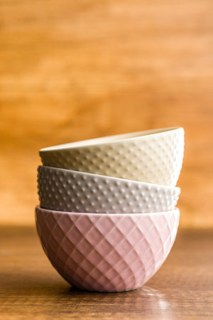 A close-up photo of three ceramic bowls stacked on a wooden tabletop. The bowls are different colors and textures, with the top bowl being a cream color, the middle bowl being a light gray with small bumps, and the bottom bowl being a light pink with a textured diamond pattern.の写真素材