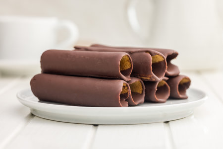 A close-up photo of chocolate-covered wafer rolls stacked on a white plate. The wafer rolls have a...