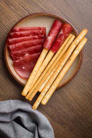 Slices of smoked meat and grissini sticks on a plate on a wooden table. Top view.