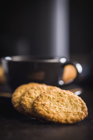 A group of oatmeal cookies is stacked neatly on a dark surface. Some cookies are placed next to the...