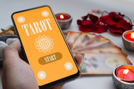 Left hand holding smartphone with tarot app, with cards and different mystical divination objects...
