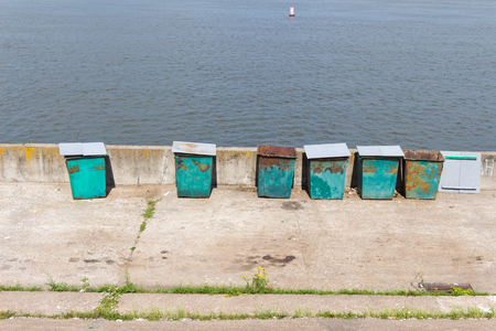 Six (6) green garbage containers standing on the stone embankment. In the background is the water and the buoy. Trash cans are old and rusty.の写真素材