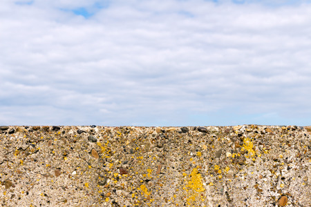 Stone (concrete) fence (block) close-up. Old and not smooth. On the surface growing moss. The background is blurred (sky and clouds).の写真素材