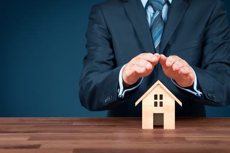 Property (family house) insurance protection concept. Insurance agent with wooden model of the house and protective gesture.の写真素材
