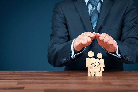 Family life insurance, services and supporting families concepts. Businessman with protective gesture and wooden figurines representing young insured family.の写真素材