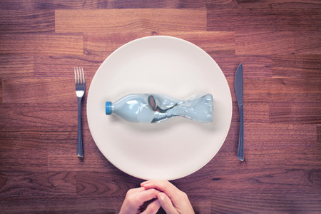 Microplastics in food concept. Microplastics and plastic pollution are global environmental problem. PET (plastic) bottle in shape of fish on plate. Knife and fork beside plate.の写真素材
