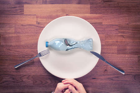 Microplastics in food concept. Microplastics and plastic pollution are global environmental problem. PET (plastic) bottle in shape of fish on plate. Knife and fork prepared for eat.の写真素材