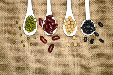 Assortment of beans in white spoon on hemp sack background. Mung beans, red beans, soy beans, black...